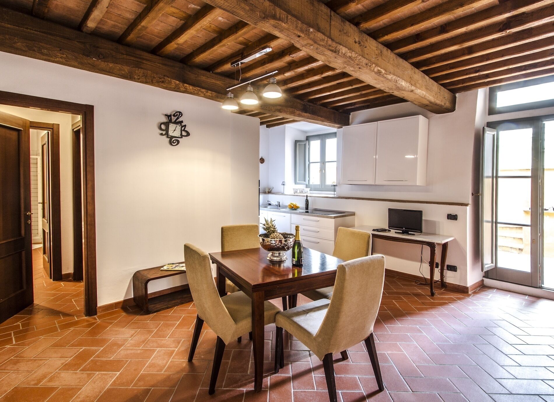A dining area with a wooden table and chairs, a kitchen in the background, and a large window.