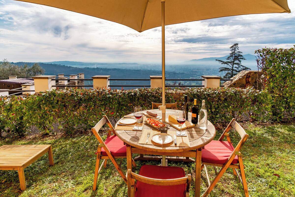 A round wooden table with chairs, a table umbrella, and a bottle of wine.