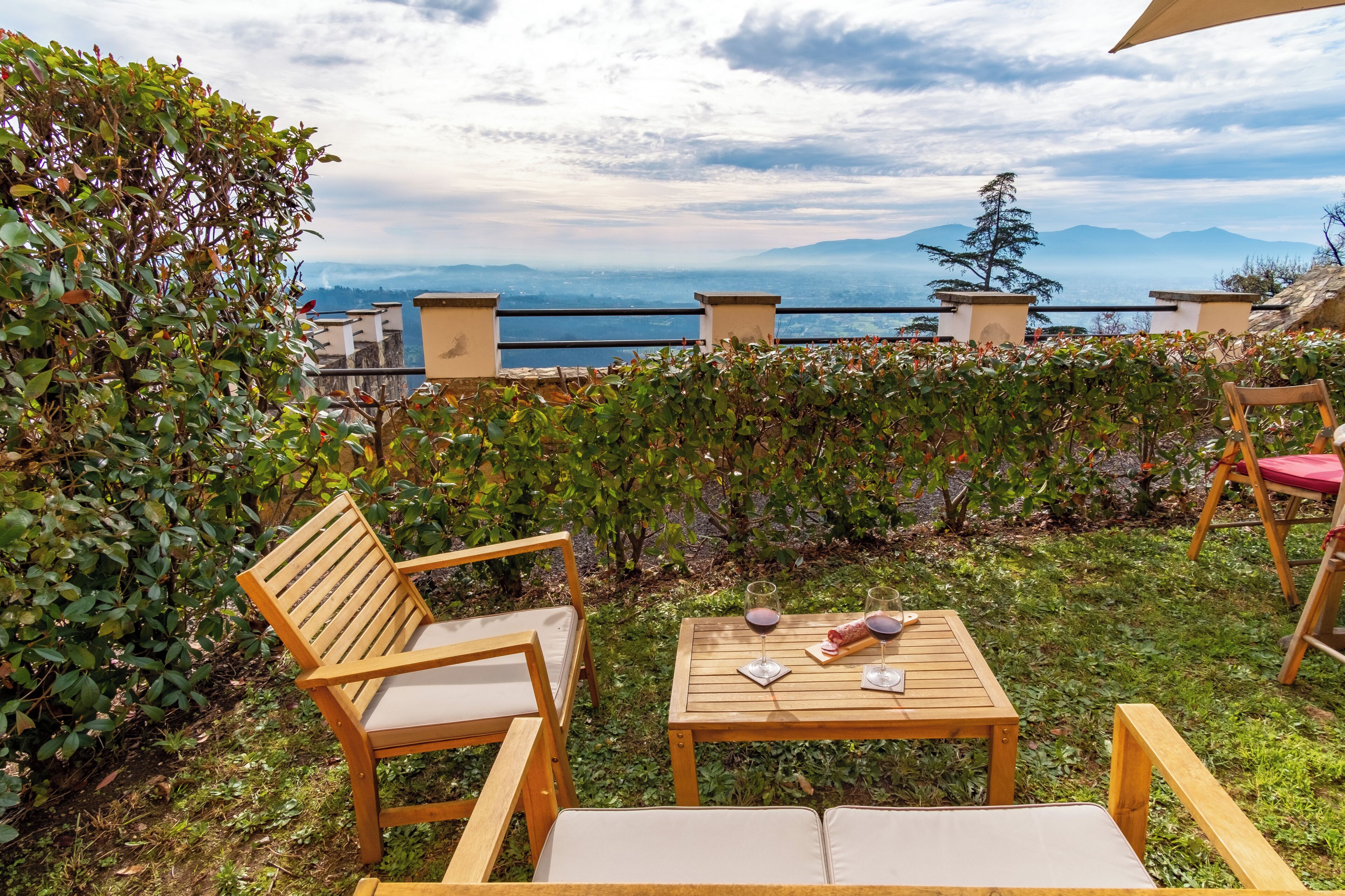 A patio with wooden furniture, a table with glasses and a napkin, and a view of the sea and mountains.