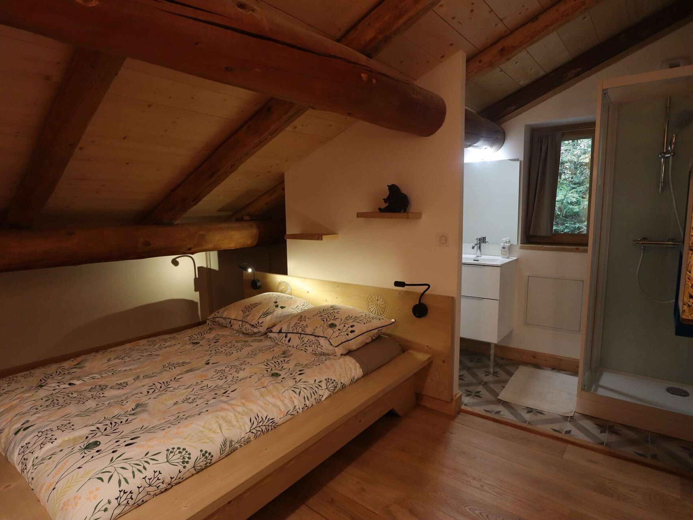 Una habitación acogedora con una cama de madera, una pequeña estantería con un objeto decorativo, una lámpara de noche y vistas a un baño con ducha.