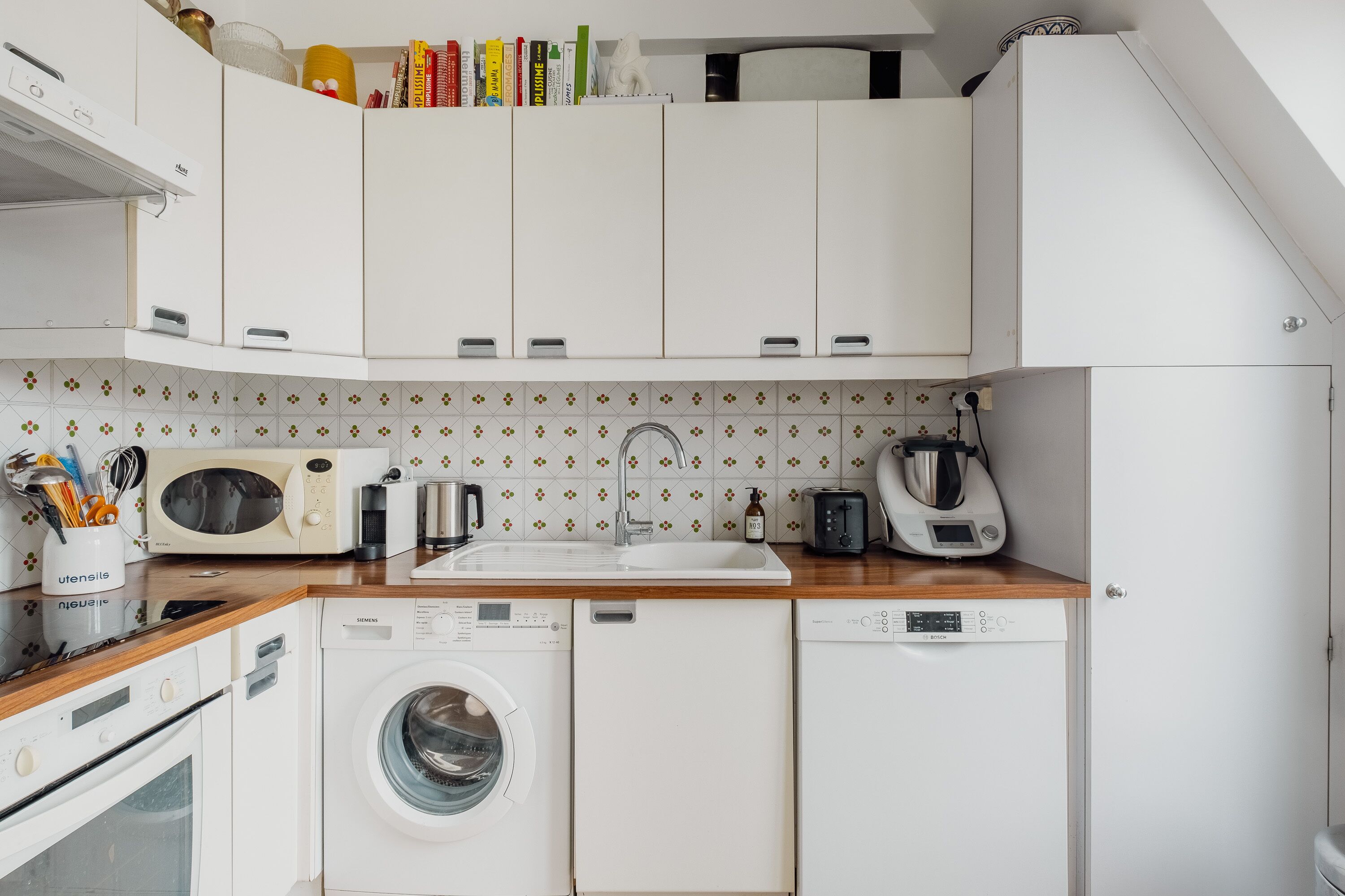Une cuisine moderne avec des meubles blancs, un lave-linge, un lave-vaisselle, un four à micro-ondes et une machine à café.