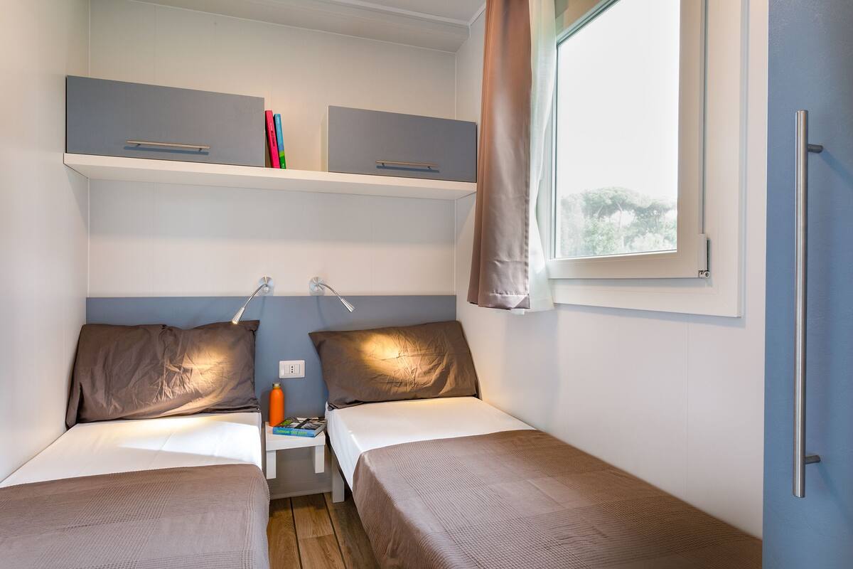 A small, well-lit room with two beds, a shelf above, and a window with curtains.