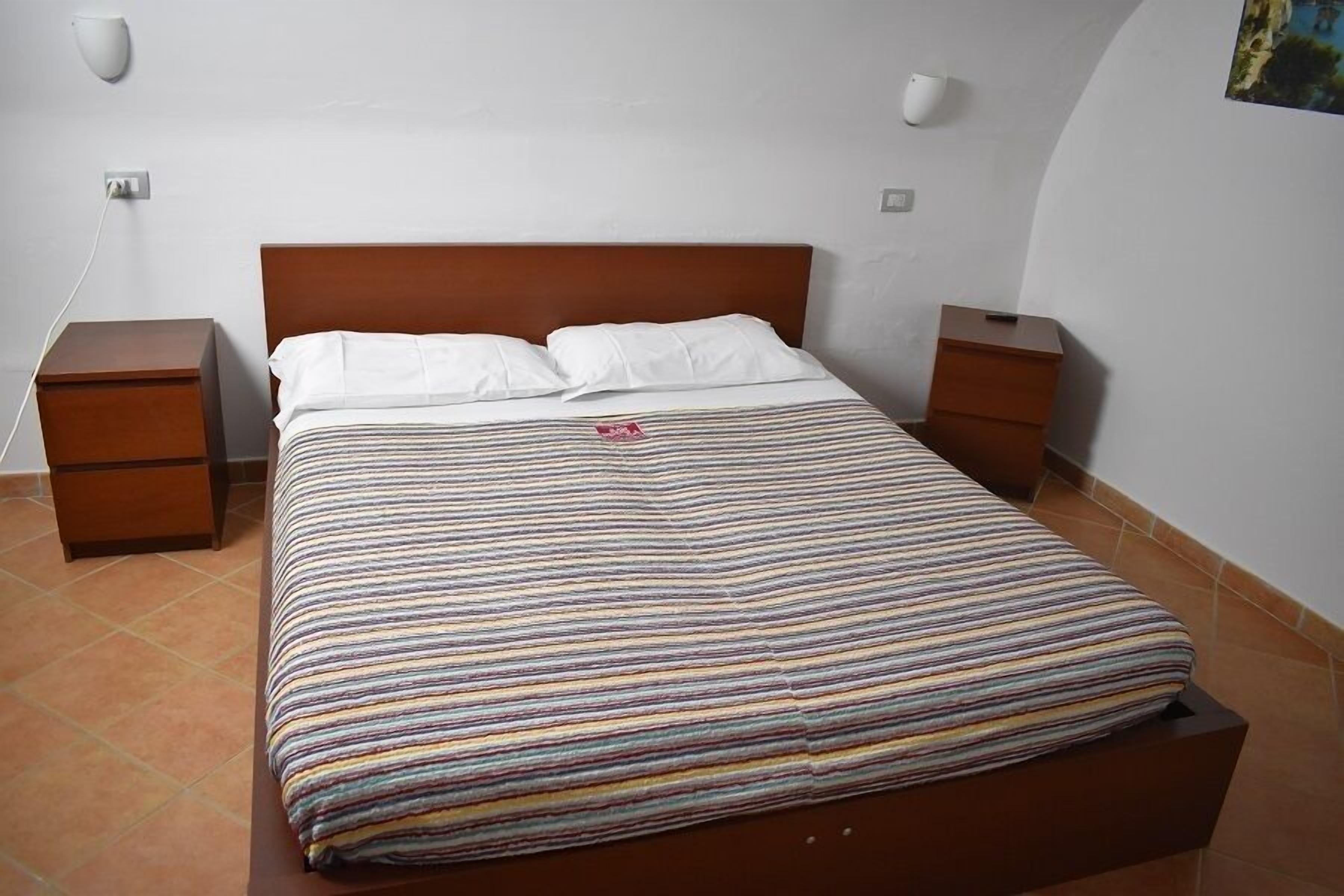 Ein ordentlich bezogenes Bett mit gestreifter Bettdecke, zwei Nachttische und einer Wandleuchte.