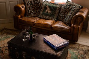 A brown leather sofa with patterned cushions, a vintage trunk with Monopoly board games and glasses, and a bottle of wine on a patterned rug.