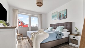 A modern bedroom with a large bed, a nightstand, a dresser, and a view of a red building outside. - Stilvolle Ferienwohnung mit Sauna, Kamin, Terrasse und Wassernähe (Büsum)