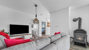 A modern living room with a grey sofa, red cushions, a flat-screen TV, a hanging light fixture, and a wood-burning stove. - Stilvolle Ferienwohnung mit Sauna, Kamin, Terrasse und Wassernähe (Büsum)