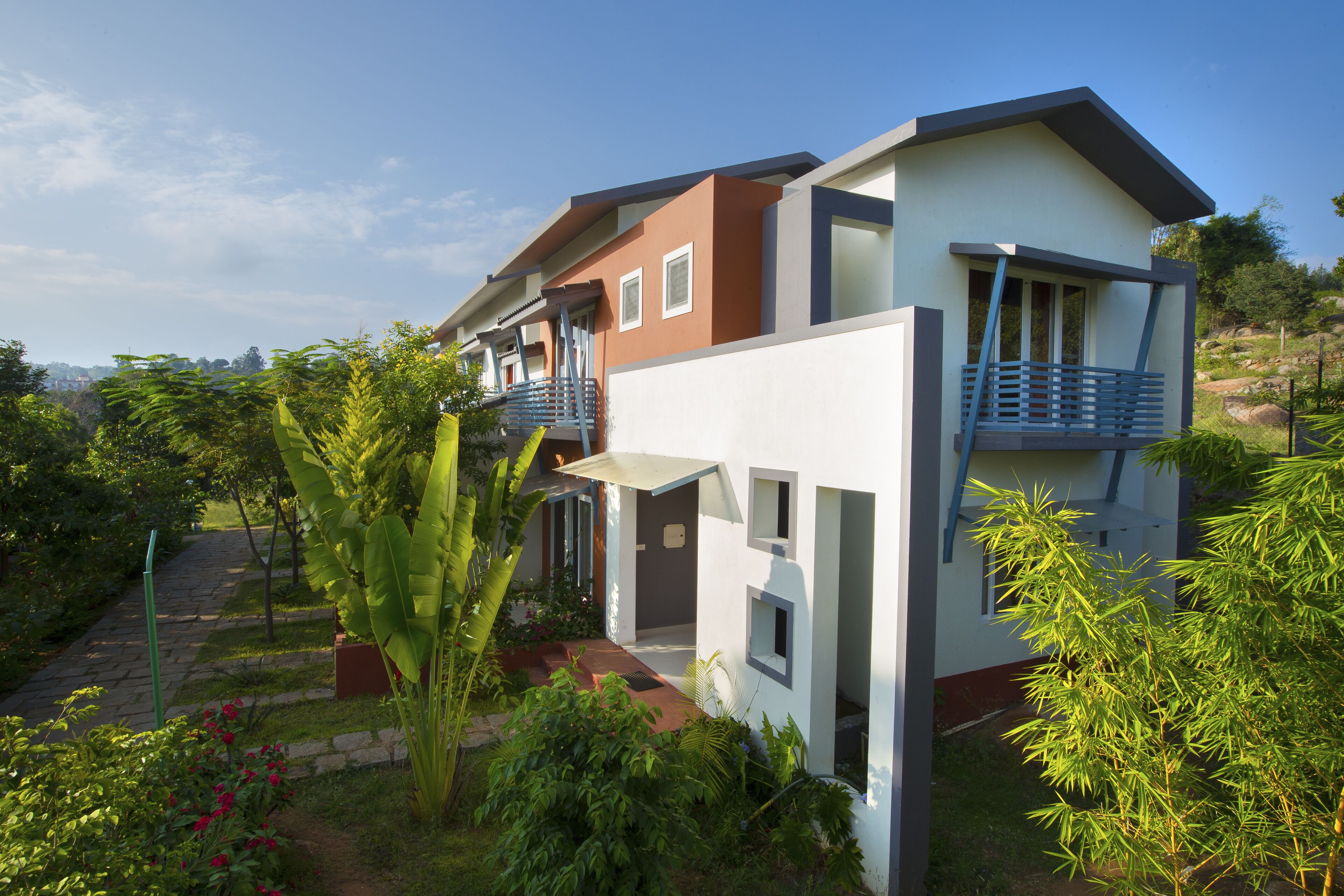 Et moderne hus med to etager og en balkon, omgivet af frodig vegetation og en stentrappe.
