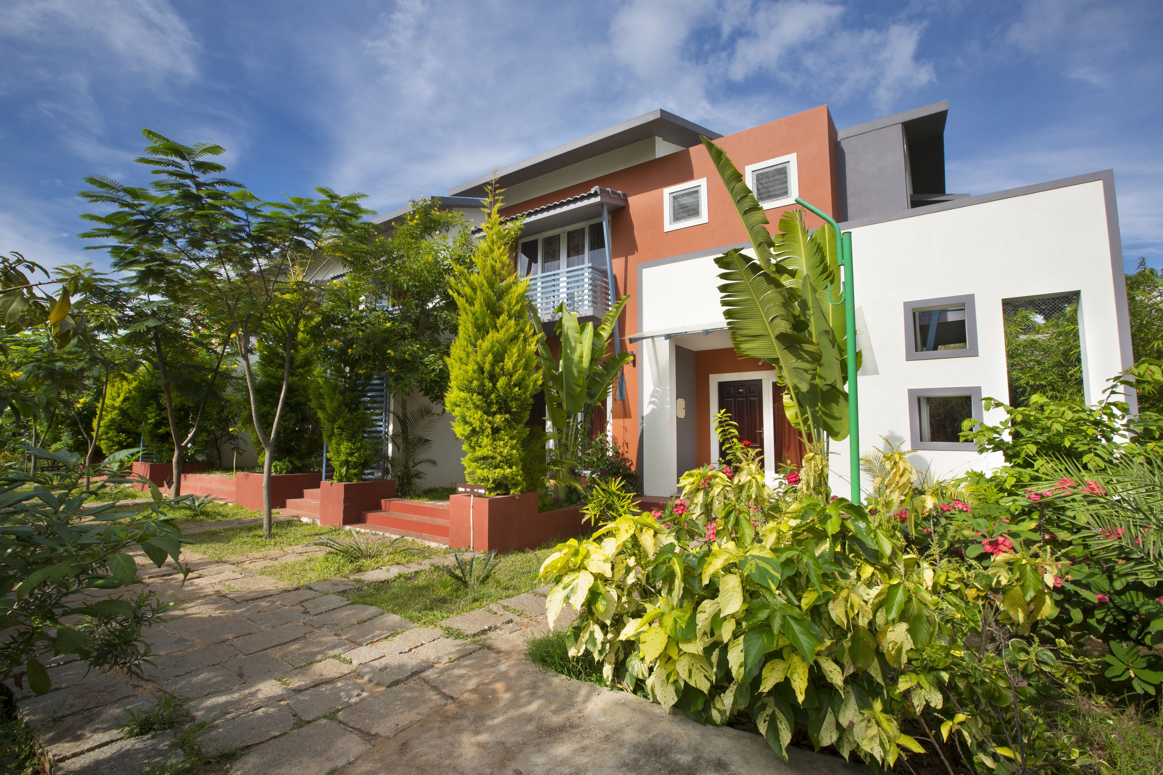 A modern two-story house with a balcony, surrounded by lush greenery and a stone pathway.