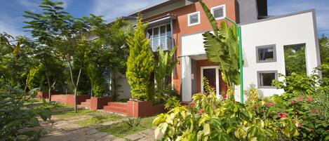 A modern two-story house with a balcony, surrounded by lush greenery and a stone pathway.