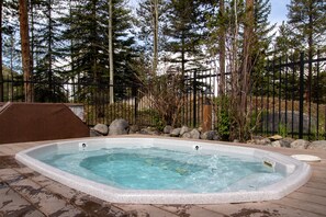 A hot tub surrounded by a wooden deck, with a metal fence and tall trees in the background.