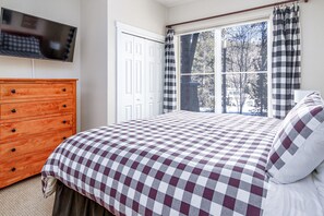 A bedroom with a checkered bedspread, a wooden dresser, a television on the wall, and a window with curtains.