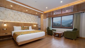 A modern hotel room with a wooden floor, a large bed with a floral bedspread, two armchairs, a small table, and a large window offering a mountain view. - SUMMIT GRAND RESORT & SPA , GANGTOK  (Gangtok)