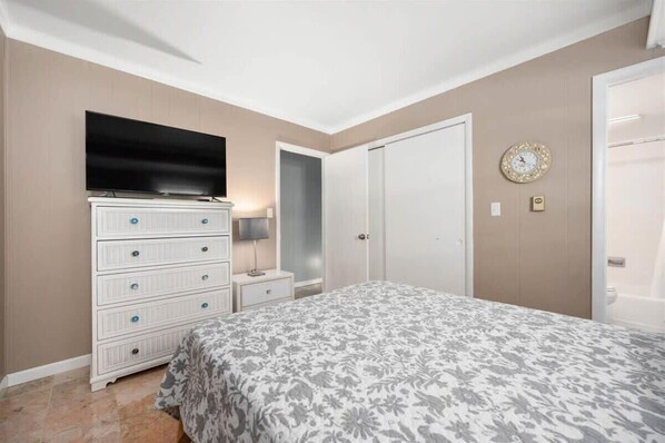 Un dormitorio con cama, cómoda, televisor, puerta y un reloj en la pared.