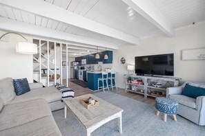 Un soggiorno moderno con divano, tavolino da caffè e poggiapiedi, una zona cucina con mobili blu e un televisore a parete.