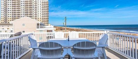 A deck with two white Adirondack chairs and a table, overlooking a beach with a high-rise building in the background.