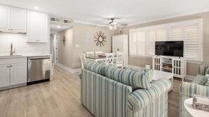 A modern kitchen with white cabinets and a dishwasher, a dining area with striped sofas and a TV, and a ceiling fan. - Three Bedroom Oceanside Condo Steps from the Beach (Ocean City)