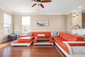A living room with a sectional sofa, a ceiling fan, a dining area with chairs, and a kitchen area with cabinets.