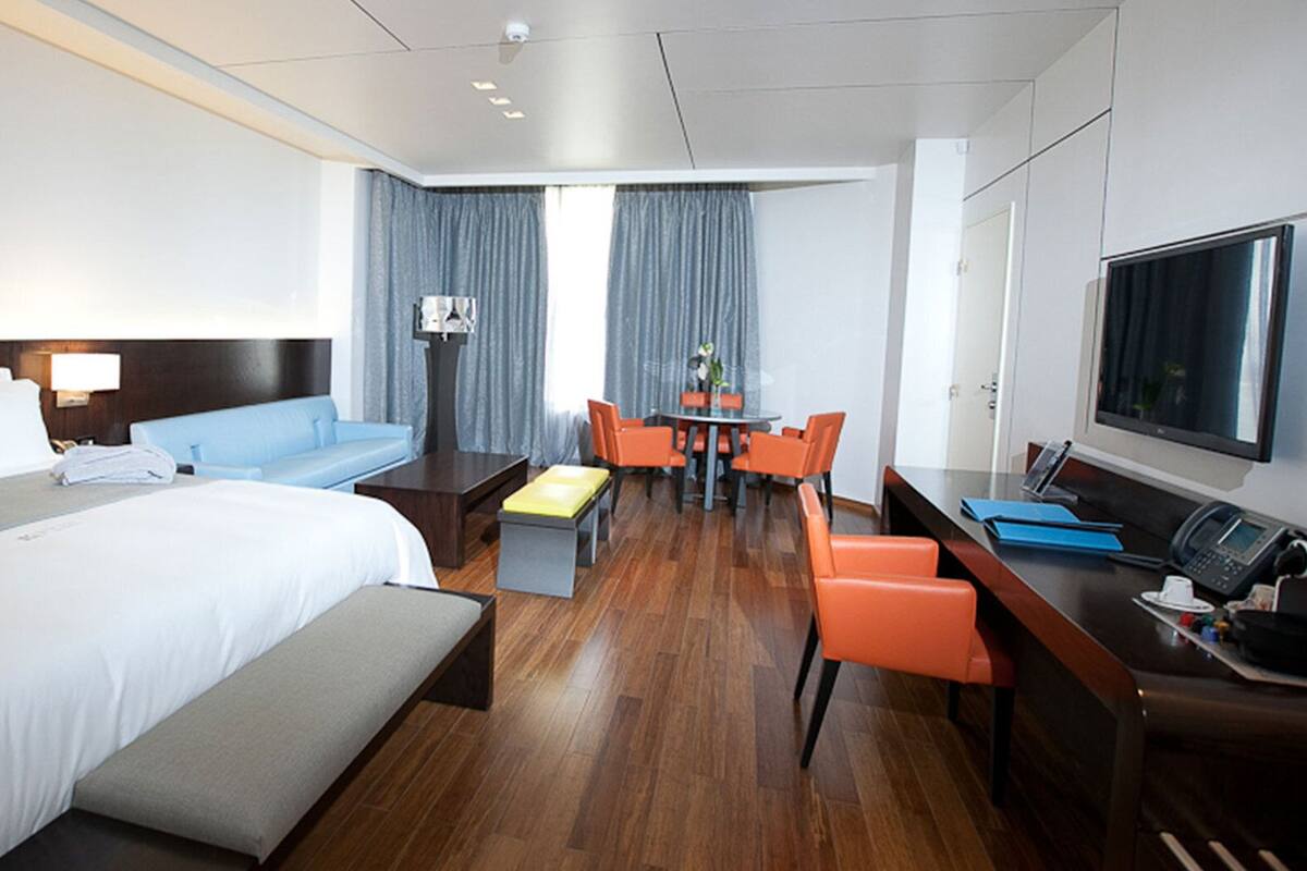A hotel room with a large bed, a desk with a computer, a TV, and a seating area with chairs.