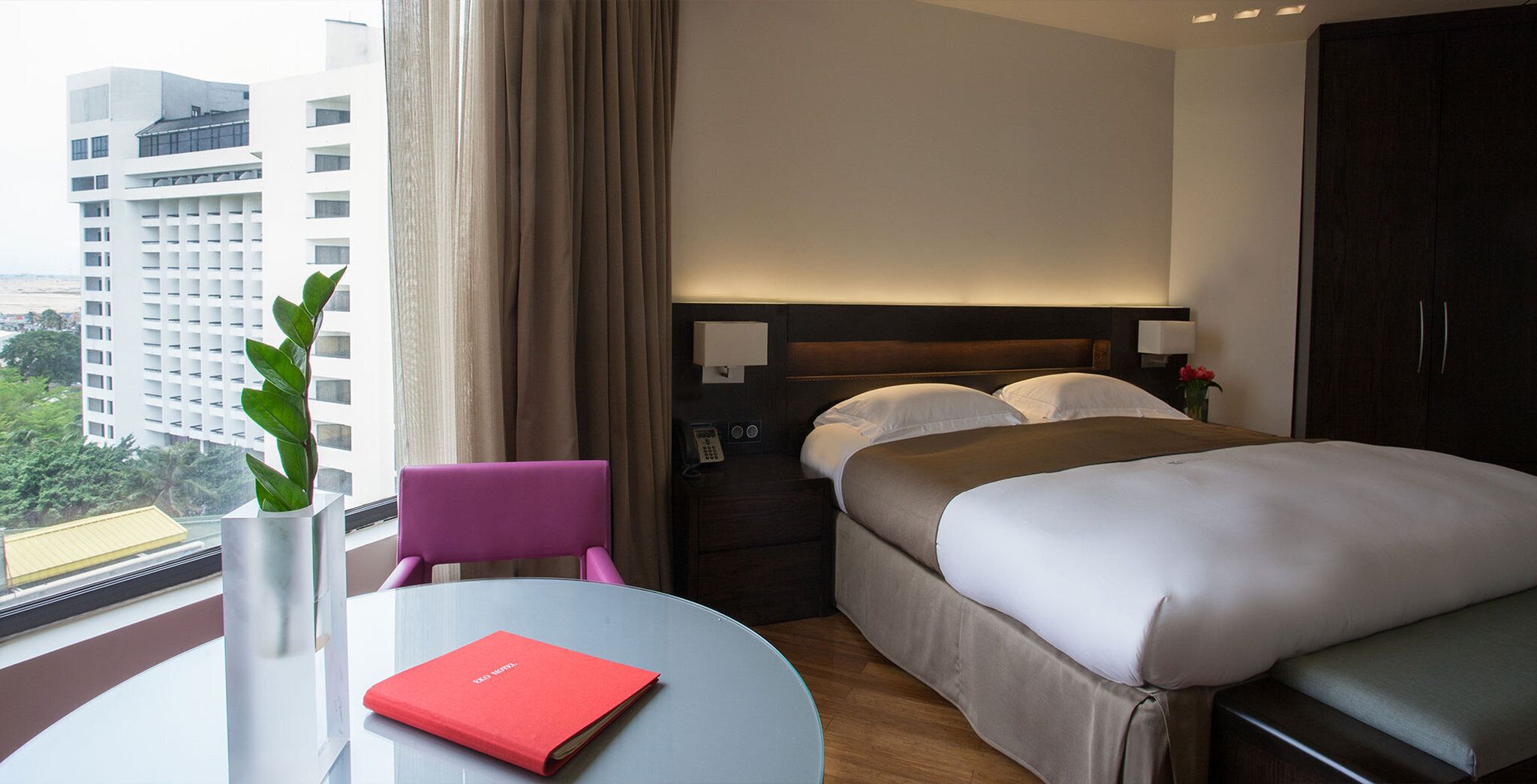 A hotel room with a bed, bedside tables, a chair, a small table, a vase with a plant, and a view of a cityscape.