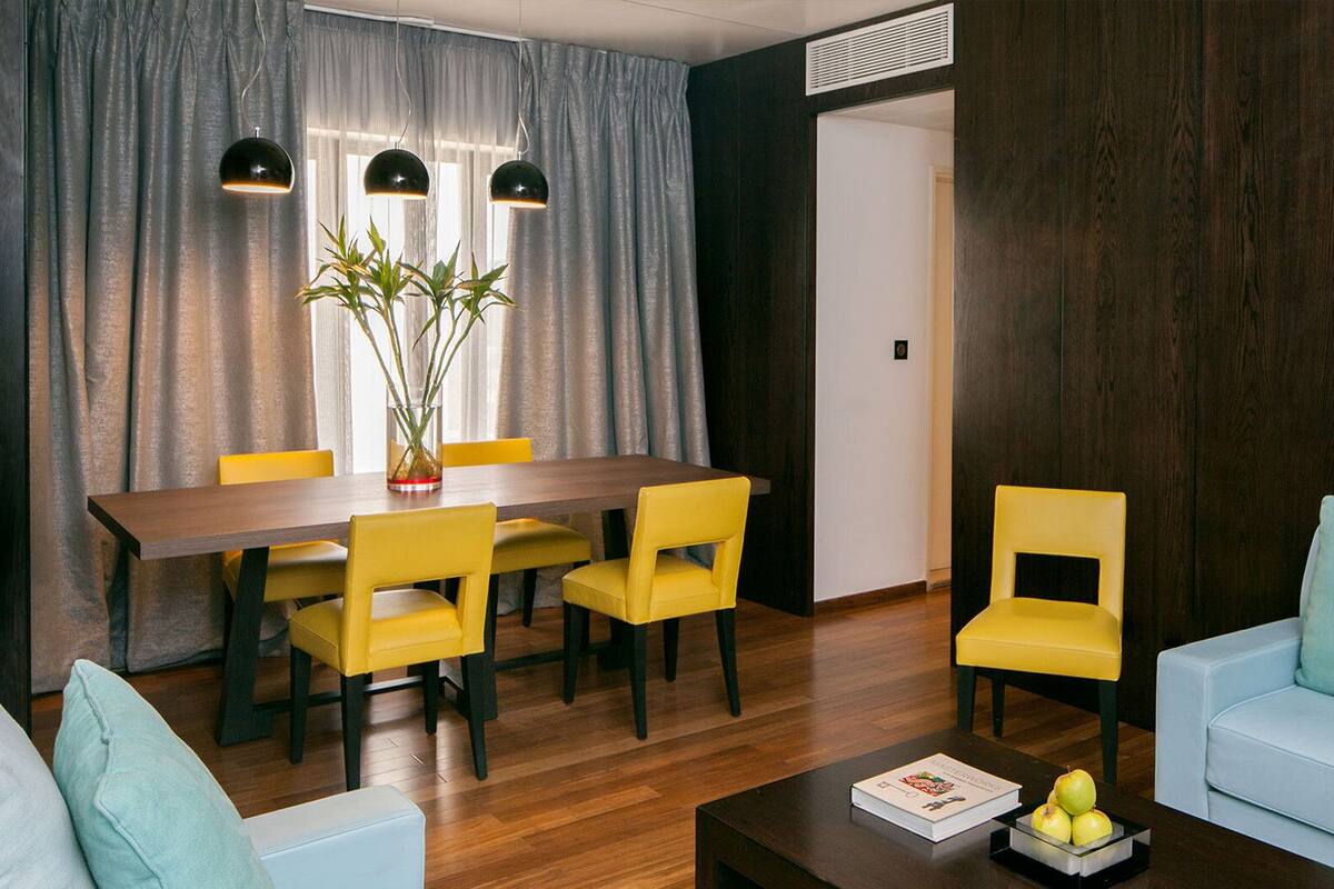 A modern living room with a dining area, featuring a wooden dining table, yellow chairs, a light blue sofa, and a coffee table with books and apples.