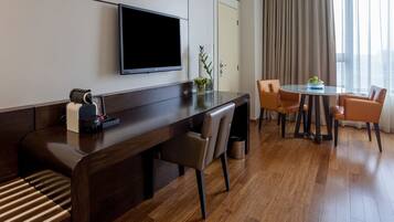 Una habitación de hotel moderna con un escritorio de madera, un televisor de pantalla plana, una zona de estar con sofá y sillones, una mesita con un jarrón, y vistas al exterior a través de amplias ventanas.