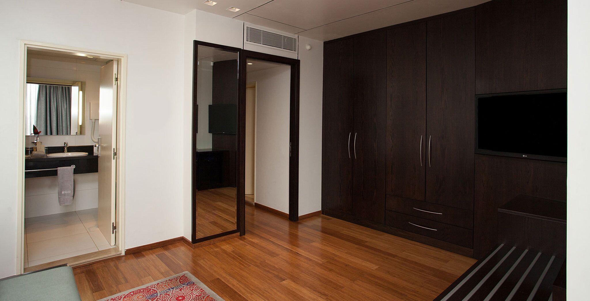 A modern bedroom with a wooden floor, a large wardrobe, a flat-screen TV, and a bathroom with a sink and mirror.