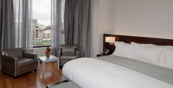 Quarto de hotel com uma cama grande, duas poltronas, uma mesa pequena, um abajur e vista da cidade através de cortinas translúcidas.