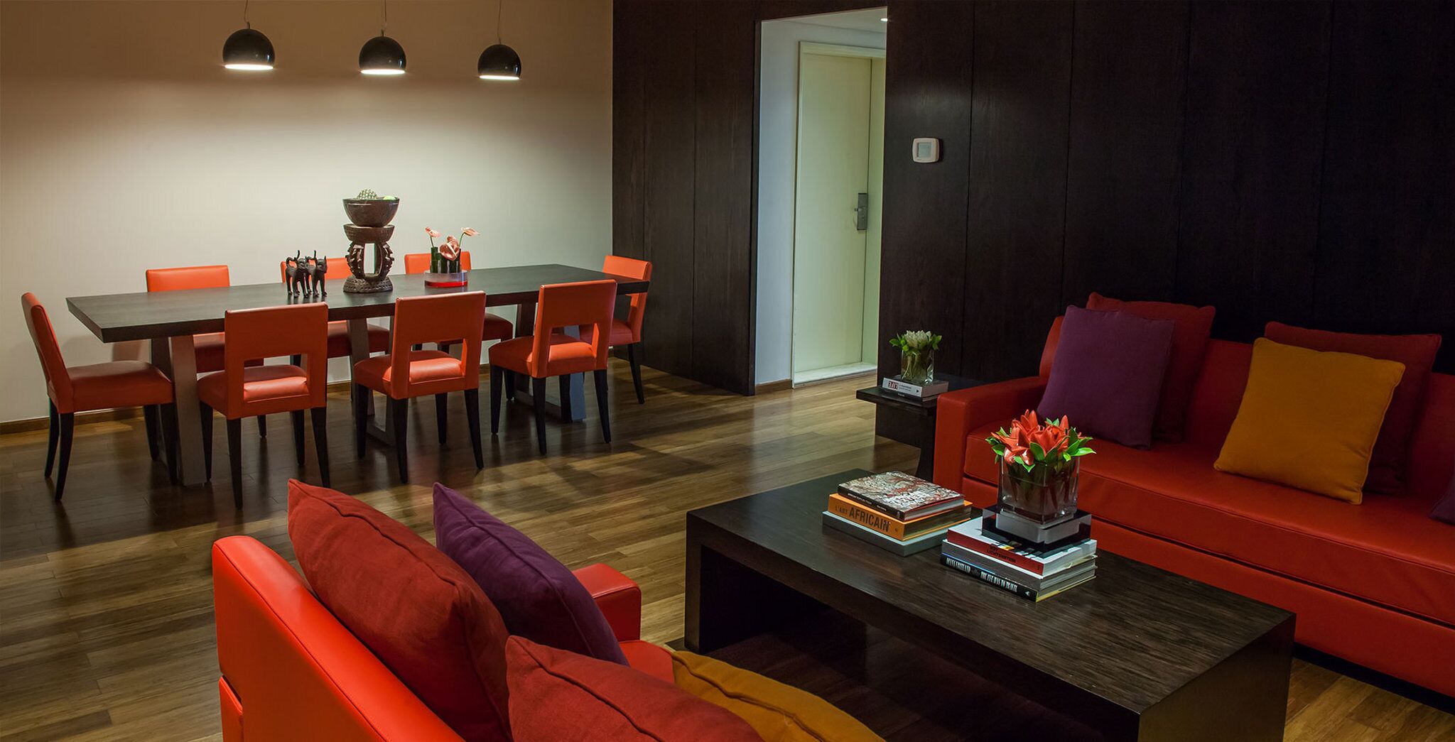 A modern living room with a red sofa, orange chairs, a coffee table with books, and a dining area with a table and chairs.