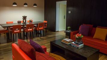 A modern living room with a red sofa, orange chairs, a coffee table with books, and a dining area with a table and chairs.