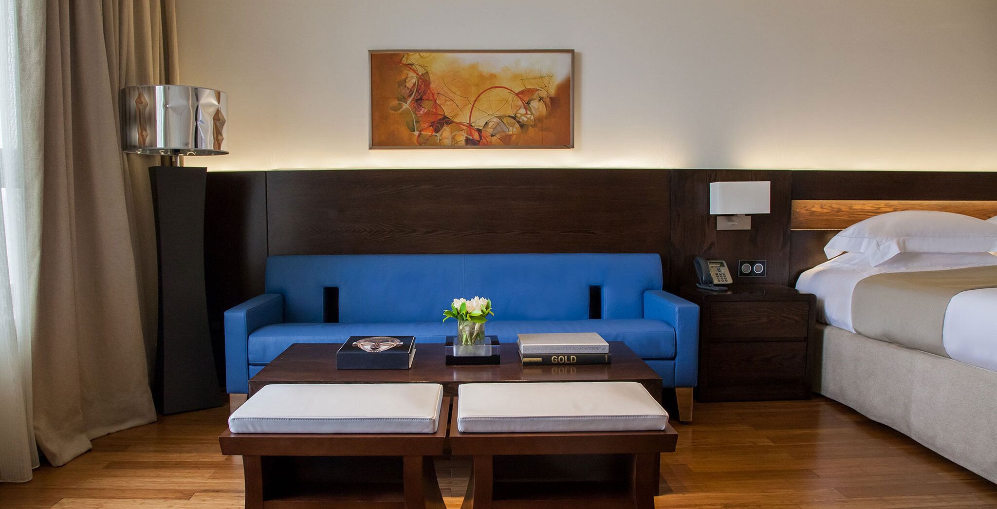 A hotel room with a blue sofa, a wooden coffee table with a vase of flowers, a bedside table with a phone, and a bed with white linens.