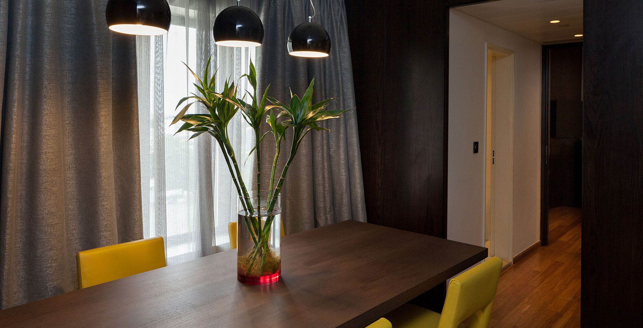 A dining area with a wooden table, yellow chairs, a glass vase with a plant, and black pendant lights.
