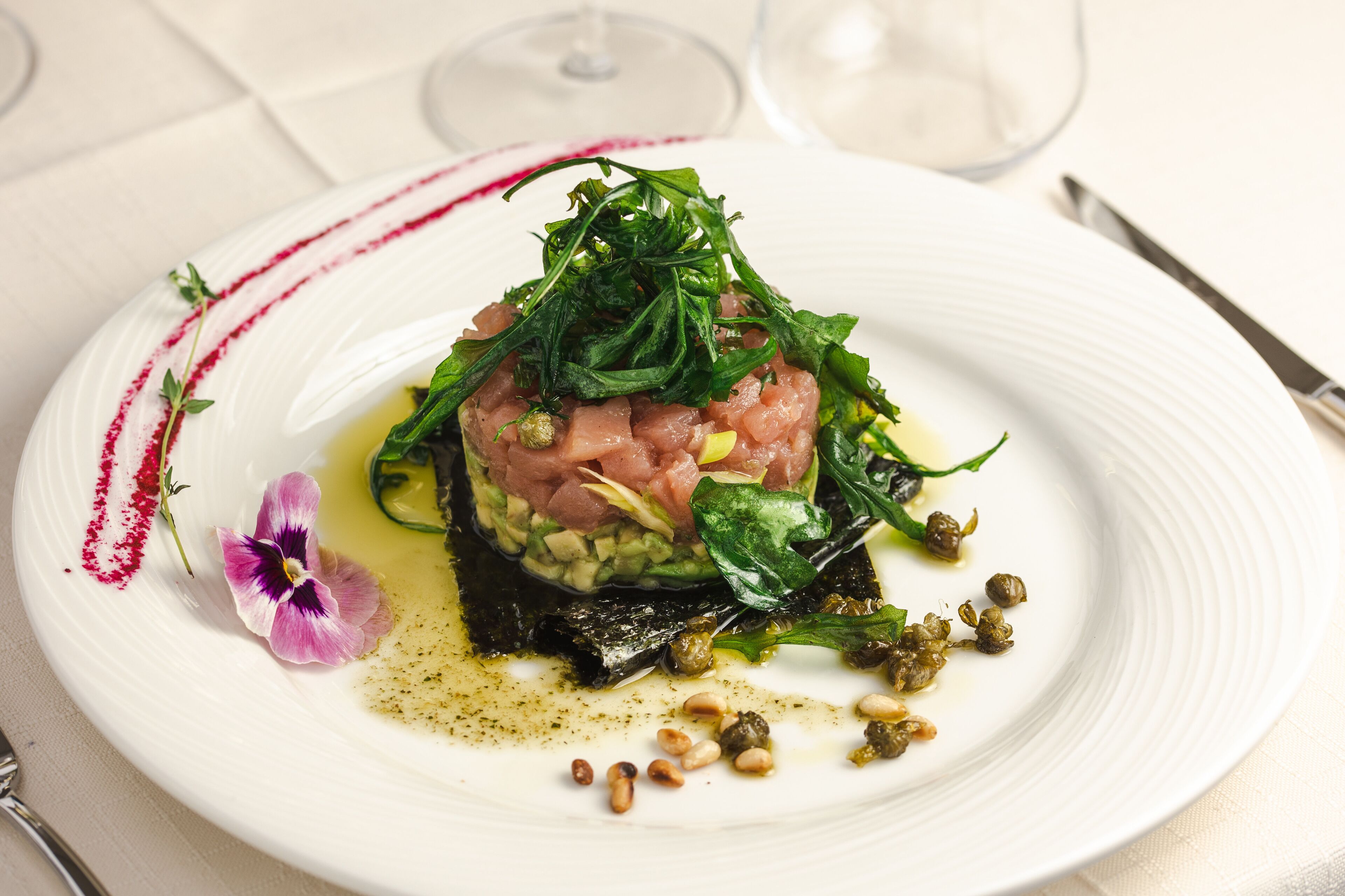 A white plate with a dish of raw fish, greens, and capers, garnished with a flower and a red sauce line.