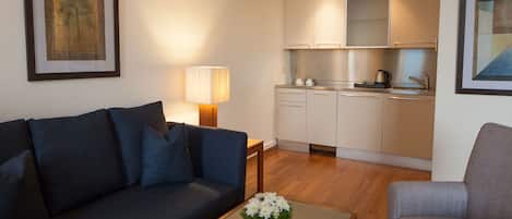 A living room with a dark blue sofa, a grey armchair, a small table with a vase of flowers, a lamp, and a kitchen area with white cabinets and a coffee maker.