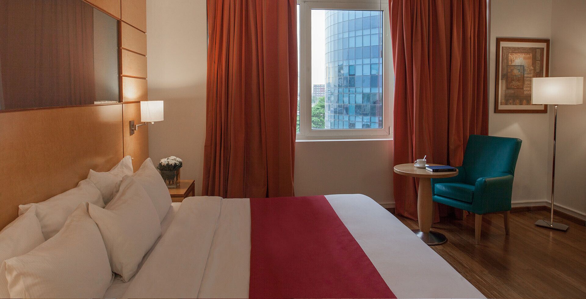 A hotel room with a bed, a chair, a small table, a lamp, and a window with curtains.