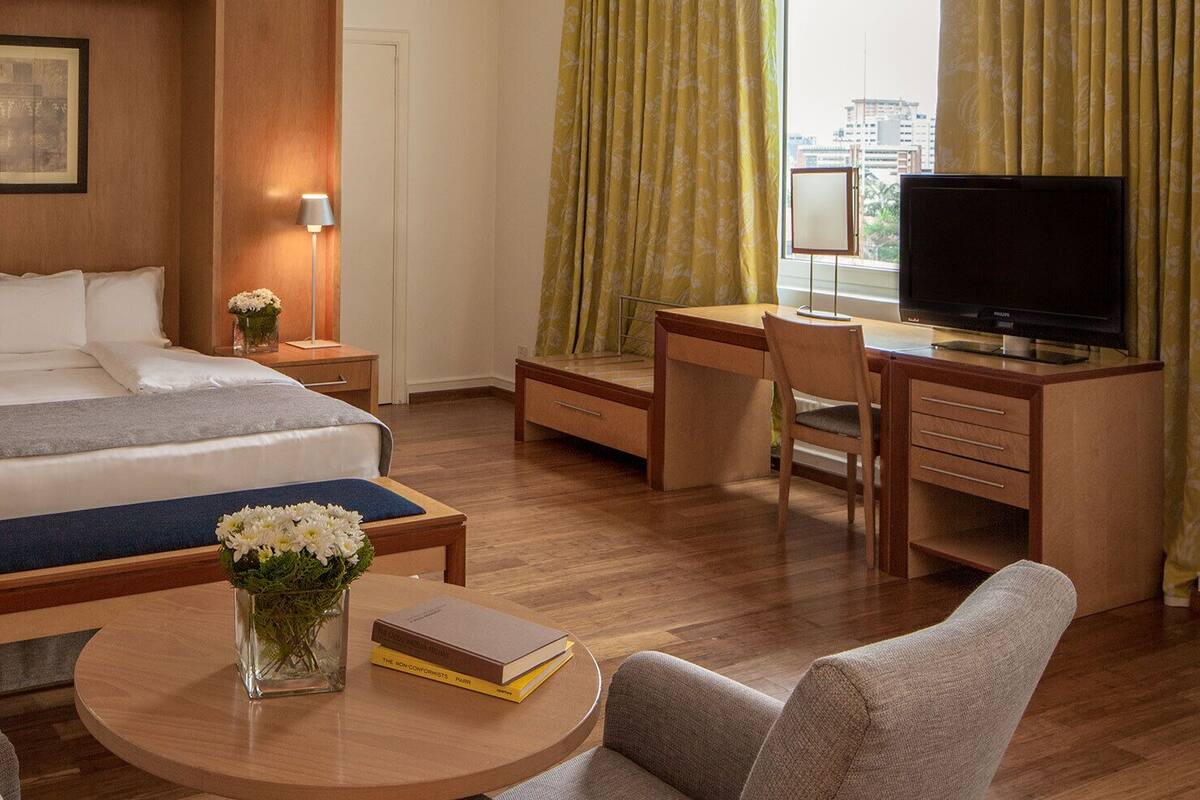 A hotel room with a bed, bedside tables, a desk, a chair, a TV, and a window with curtains.