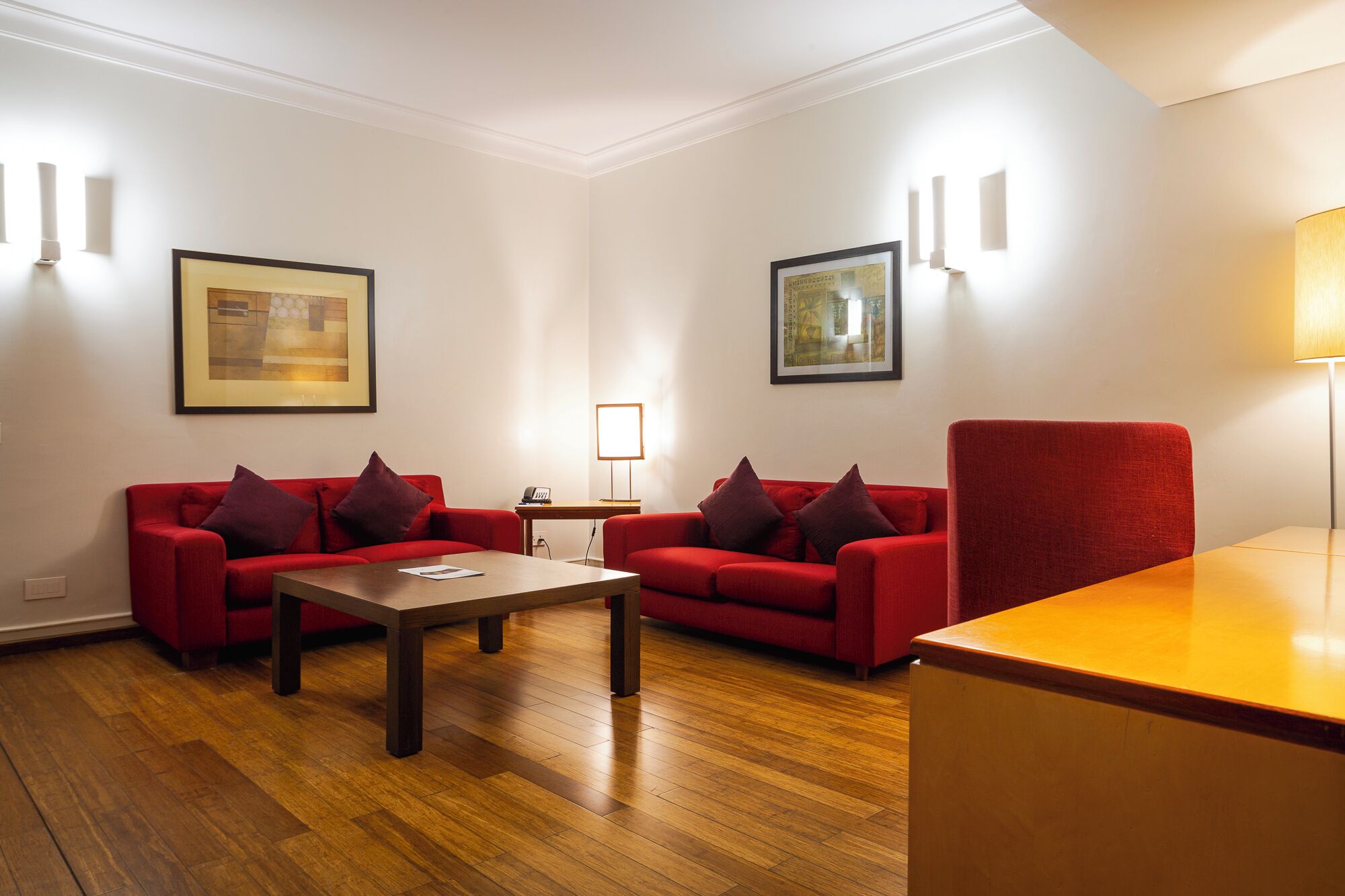 Een kamer met twee rode banken, een houten salontafel, een klein bijzettafeltje en een rode stoel. Er hangen twee kunstwerken in lijsten aan de muren en er staat een lamp aan de rechterkant.