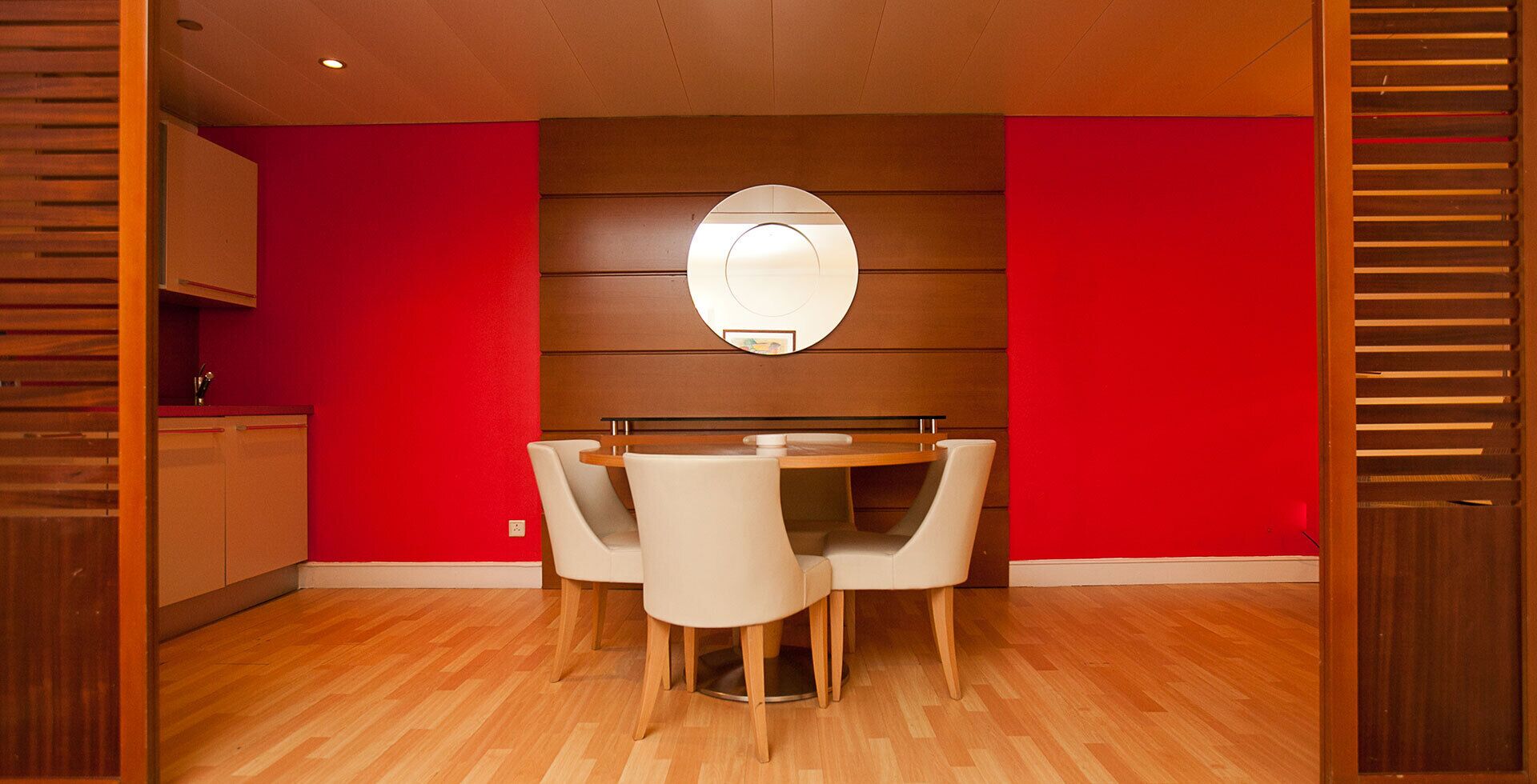 A dining area with a wooden table and four chairs, a red wall, and a wooden cabinet.