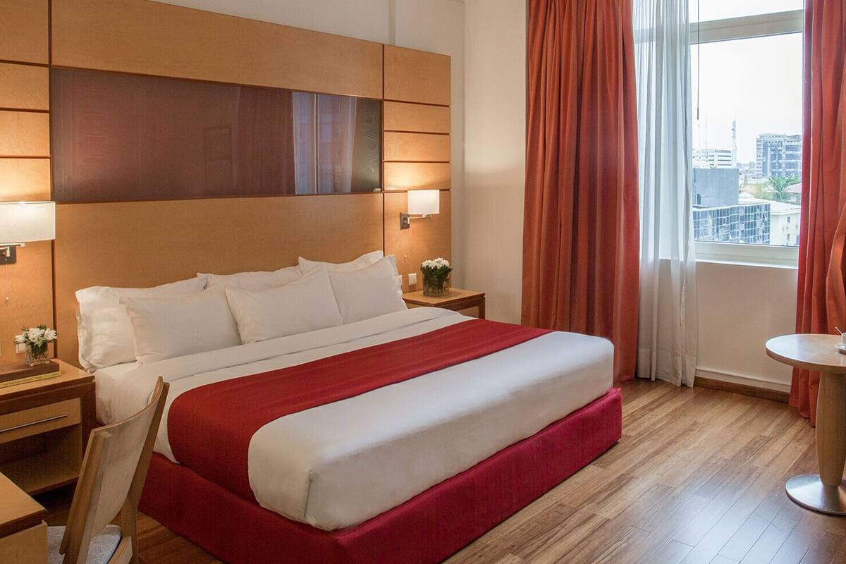 A hotel room with a large bed, two bedside tables, a small table with a cup, a chair, and a view of the city through the window.