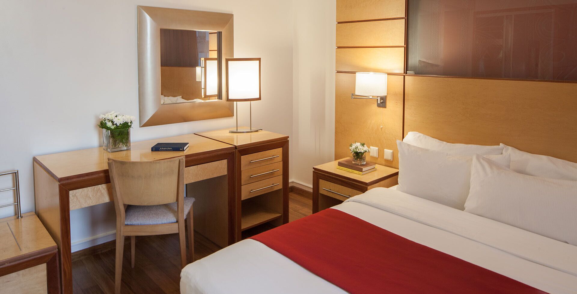 Quarto de hotel com cama, escrivaninha, cadeira, mesa de cabeceira, espelho e luminária na parede.