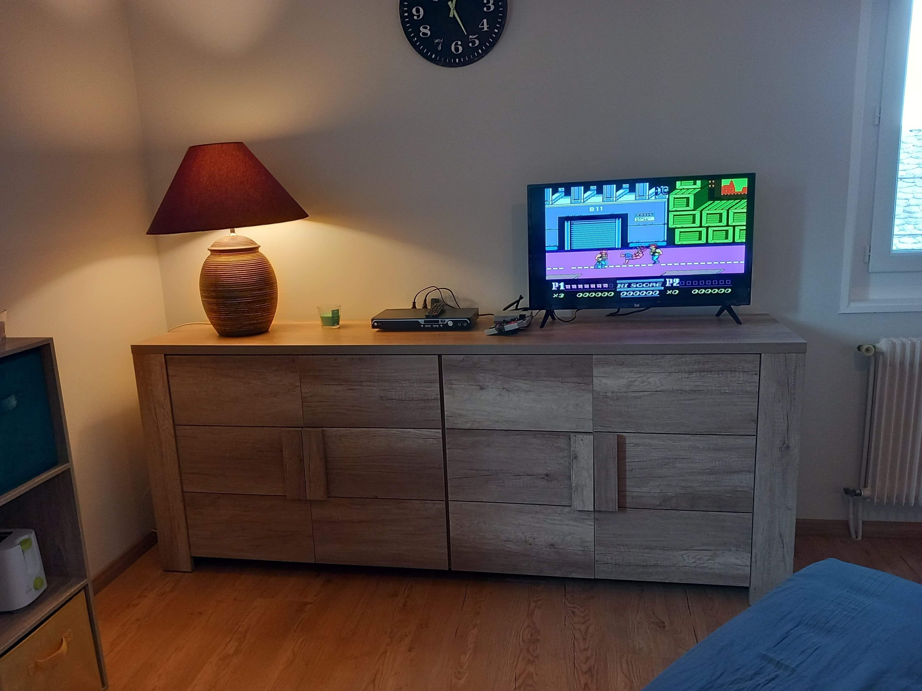 Un meuble en bois sur lequel est posée une télévision diffusant un jeu vidéo, une horloge accrochée au mur et une lampe sur le dessus.