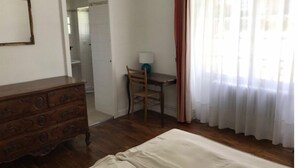 Une chambre à coucher avec une commode en bois, un lit, un bureau avec une chaise et une fenêtre avec des rideaux.