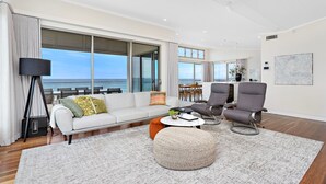 Una sala de estar moderna con un ventanal grande con vista al océano, un sofá, dos sillones, una mesa de centro y una lámpara de pie.