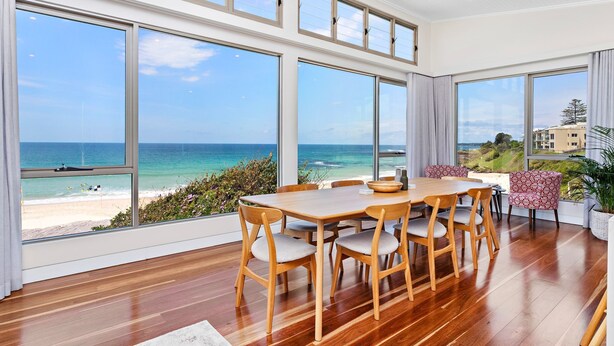 Un comedor amplio con una gran mesa de madera y sillas, ventanas grandes con vistas a la playa y al océano, y una planta en maceta a la derecha.
