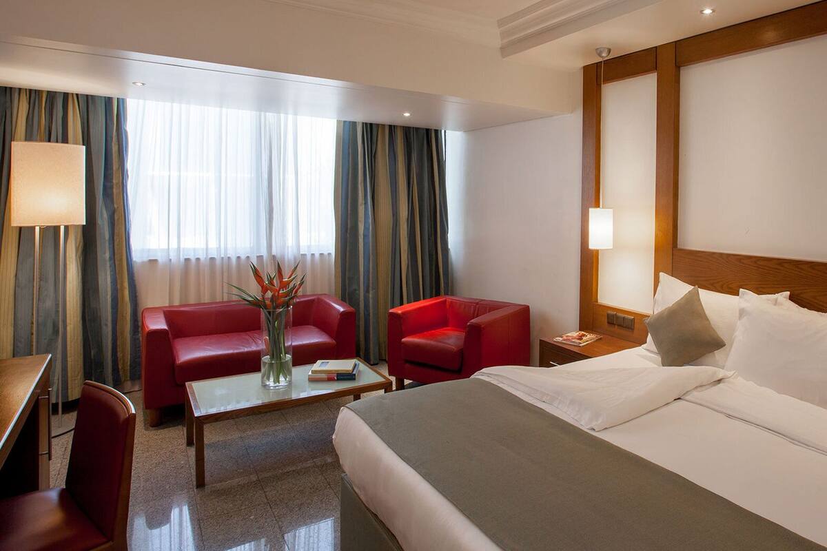 A hotel room with a bed, a desk, a red chair, a small table with a vase, and a nightstand with a lamp.