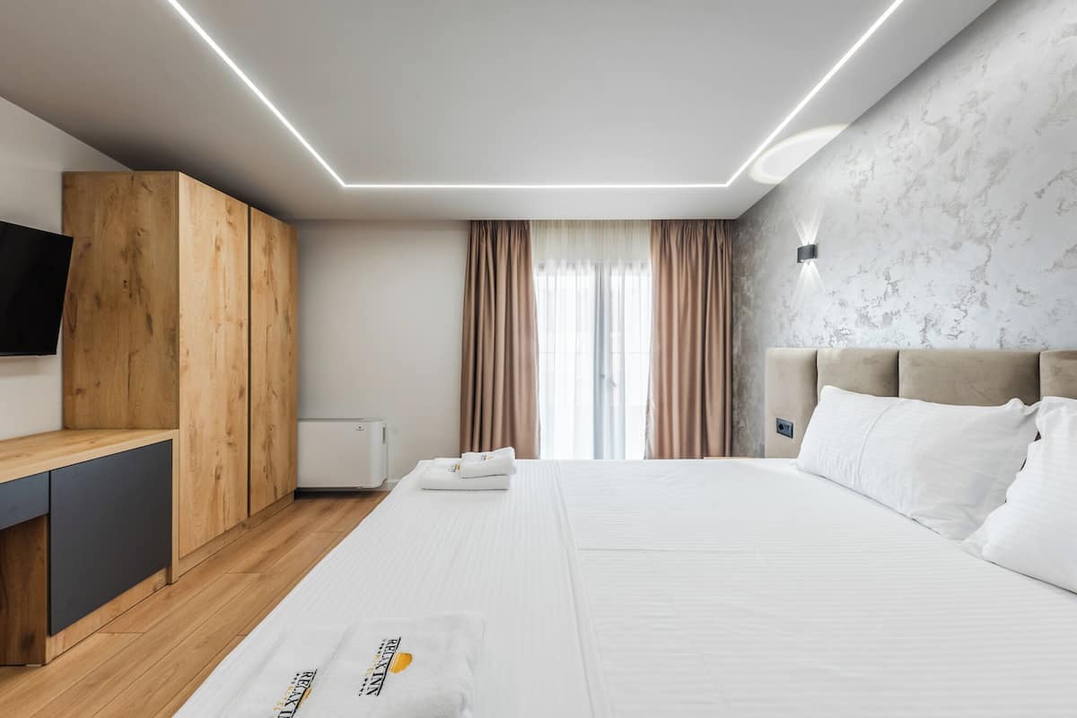 A modern hotel room with a wooden wardrobe, a flat-screen TV, a bed with white linens, and a large window with curtains.
