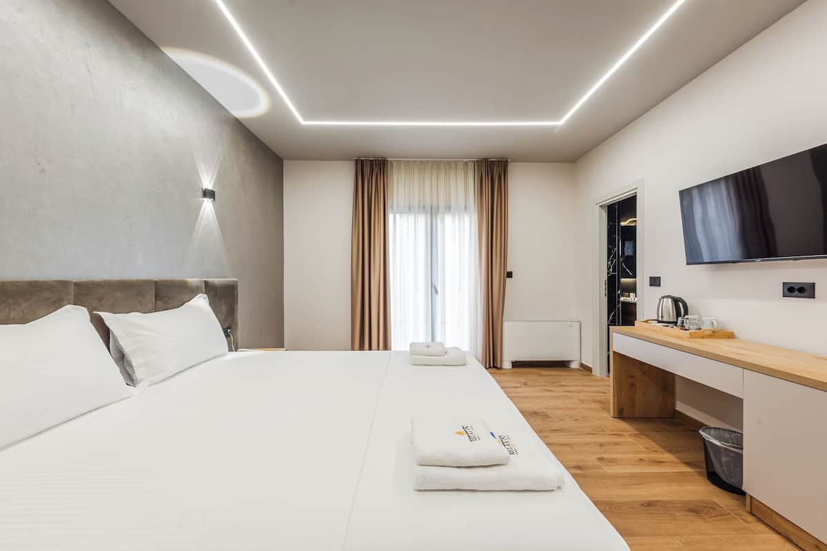 A modern hotel room with a large bed, a flat-screen TV, a wooden desk, and a view of the city.