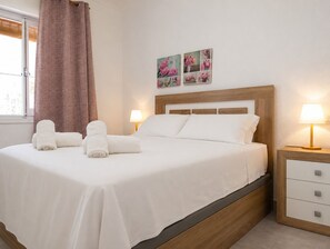 A neatly made bed with white linens and two rolled towels, a wooden headboard, bedside lamps, and a small bedside table with drawers. - Charming House with Nature (El Chaparral)