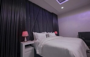A modern bedroom with a large bed, dark curtains, and a small bedside table with a lamp.