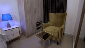 A room with a wooden wardrobe, a small bedside table with a lamp, a sofa, and a glass coffee table. - 3H VILLAS HOTEL & LUXURY SHORT-LET  (Lekki)