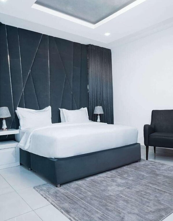 A modern bedroom with a large bed, two bedside tables with lamps, a dark upholstered armchair, and a geometric-patterned headboard.